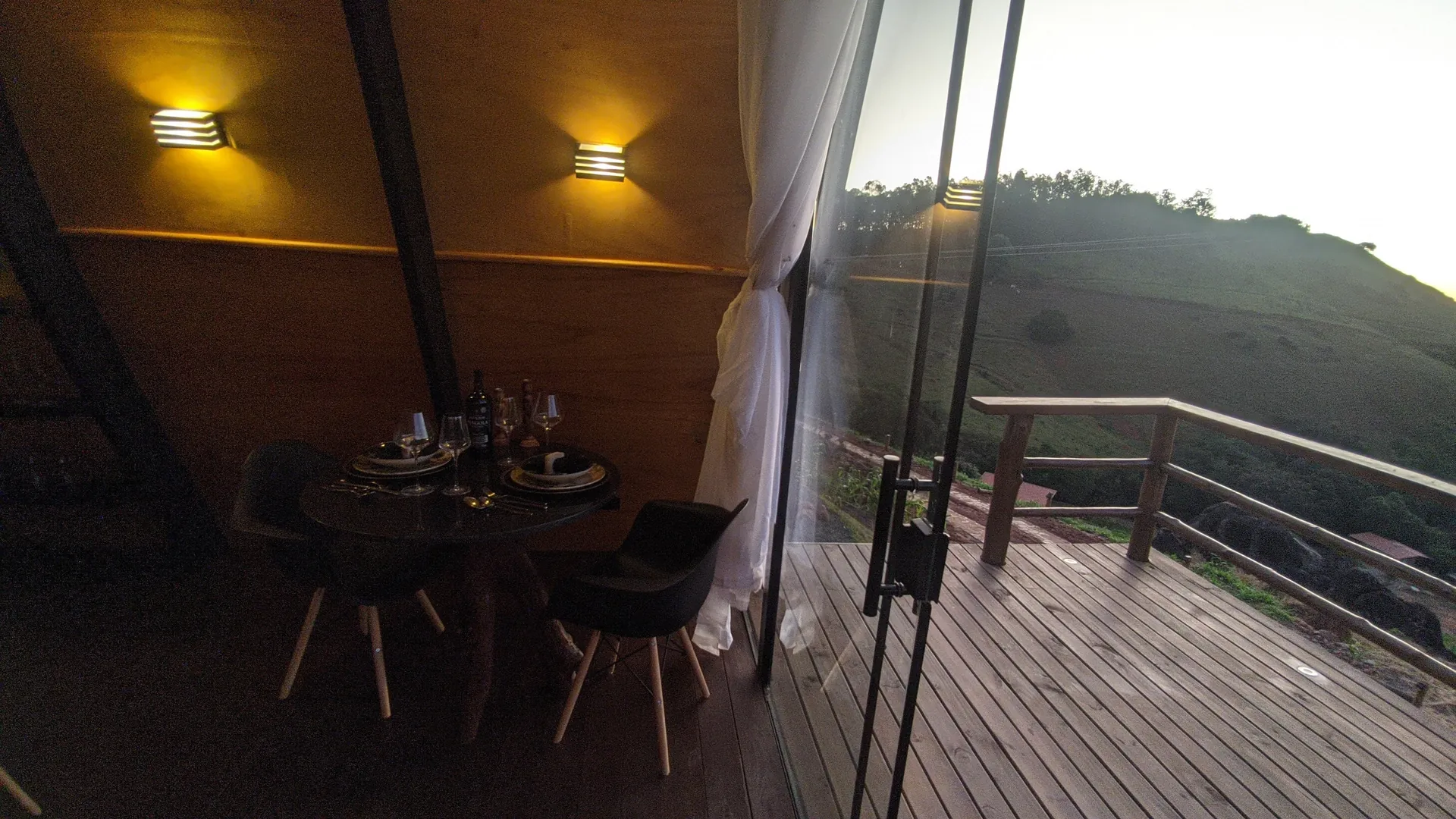 Mesa posta para dois junto ao vidro do chalé, com o deck e o vale ao entardecer